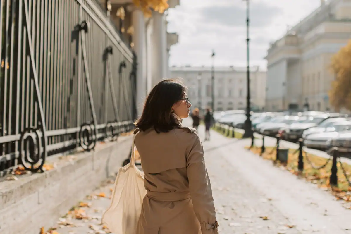 Femeie în trenci care merge pe un trotuar urban într-o zi însorită
